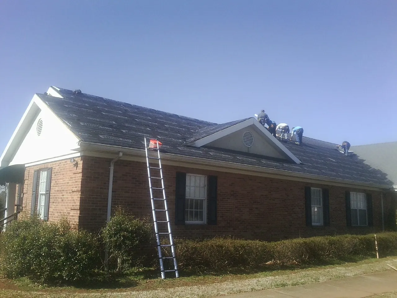 Crew tearing shingles off a brick ranch roof with a ladder set — Metal Roof Repair work by our crew in Capron
