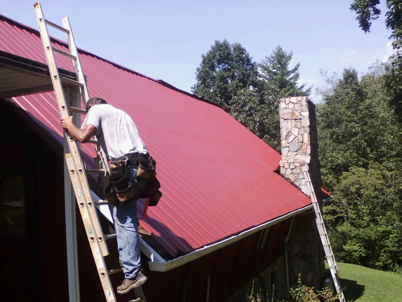 Roofer on a ladder beside a red metal roof and stone chimney — Soffit & Fascia Repair by Roofing Brothers in Capron